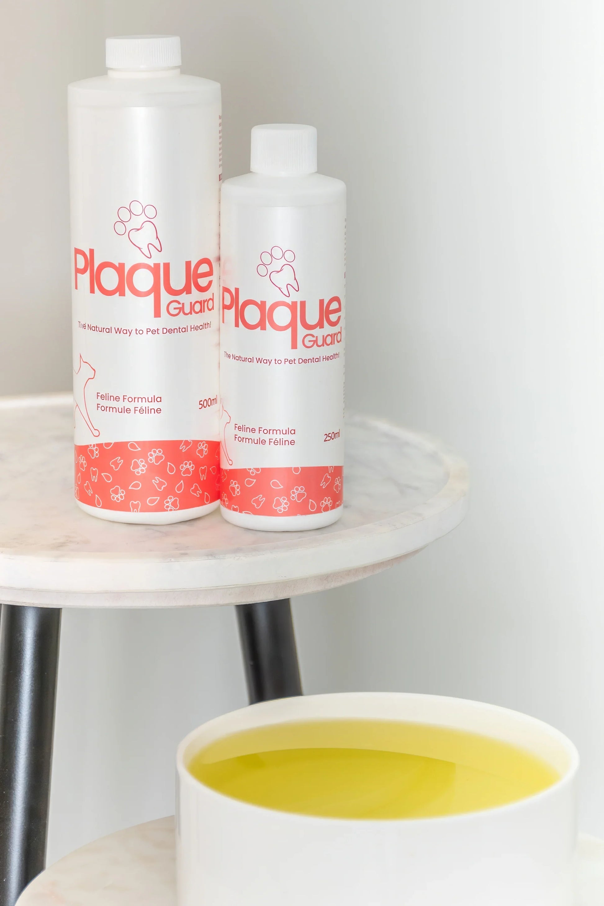 Two Plaque Guard dental water additive bottles for pets on a small marble table, next to a bowl of water.